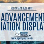 A blog post graphic from AGDisplays with the title "THE ADVANCEMENT OF AVIATION DISPLAYS" in large, bold blue letters against a slightly transparent white rectangular overlay. Above the title, smaller grey text reads "AGDISPLAYS BLOG POST". Below the title, the AGDisplays logo in blue and white is centered above "www.agdisplays.com" in smaller blue text. The background image is a slightly blurred view of a sophisticated aircraft cockpit with multiple digital displays and various controls, suggesting advanced avionics technology.