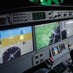The interior of an advanced aircraft cockpit is shown, dominated by several large, brightly lit digital displays integrated into the instrument panel. The primary display on the left shows flight instrumentation, including an artificial horizon, altitude, airspeed, and vertical speed indicators, along with navigational data. The central displays depict a moving map with weather radar overlay, and possibly other navigational or system information. Smaller digital and analog gauges are visible below the main screens, along with numerous buttons, knobs, and switches. A control yoke is prominent in the foreground on the right. The overall impression is of sophisticated avionics technology designed for modern flight management.