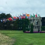 A wide shot shows the exterior of a modern, concrete building with the large, stylized letters "F" and "E" stacked vertically and connected in the center, forming a logo. To the right of this logo, the word "FUTURE" is spelled out in large, sans-serif letters. In front of the building, a well-maintained green lawn is bordered by a long row of flagpoles, each displaying a different national flag. Purple flowers are planted in a bed along the base of the "FUTURE" sign and around a few small evergreen trees. The sky is overcast and grey, suggesting a cloudy day. Trees are visible in the background behind the flags and the building.