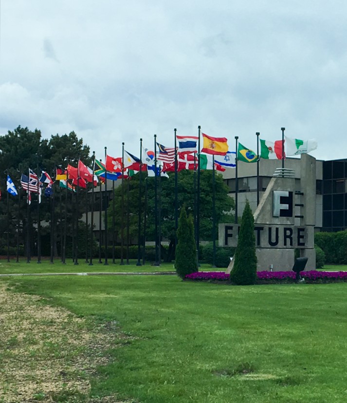 A wide shot shows the exterior of a modern, concrete building with the large, stylized letters "F" and "E" stacked vertically and connected in the center, forming a logo. To the right of this logo, the word "FUTURE" is spelled out in large, sans-serif letters. In front of the building, a well-maintained green lawn is bordered by a long row of flagpoles, each displaying a different national flag. Purple flowers are planted in a bed along the base of the "FUTURE" sign and around a few small evergreen trees. The sky is overcast and grey, suggesting a cloudy day. Trees are visible in the background behind the flags and the building.