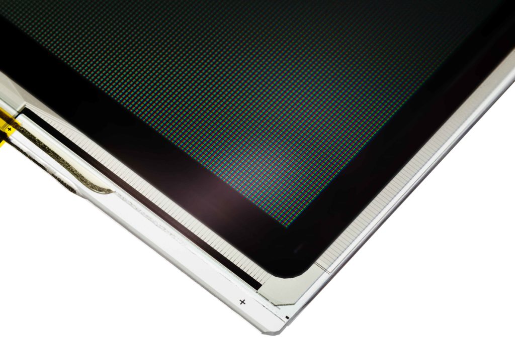 A close-up, cropped shot of the bottom-right corner of an LCD screen, showing the individual red, green, and blue pixels. The image highlights the detailed pixel grid, the black border around the display area, and the white bezel with a faint crosshair symbol at the bottom.